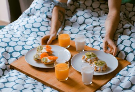 Bed Breakfast - person serving pastries on white ceramic plates with fruit juice glasses on wooden tray on top of bed