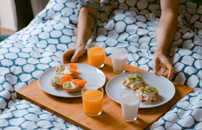 Bed Breakfast - person serving pastries on white ceramic plates with fruit juice glasses on wooden tray on top of bed